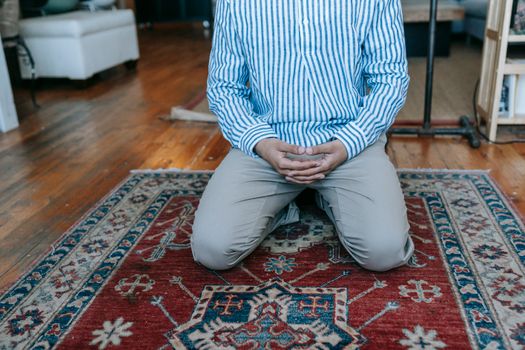 Man kneeling indoors on a patterned rug, hands clasped, in a prayerful position.