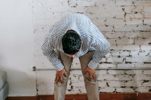 A man in casual attire bowing in a room with a white brick wall. Indoor setting.