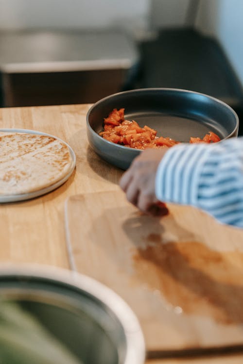 Crop unrecognizable ethnic male at table with chopping board against flatbread and cut fresh tomato in bowl at home