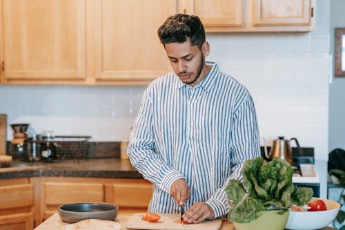 Bearded ethnic male in striped shirt cutting fresh tomato with knife against spinach leaves on table at home