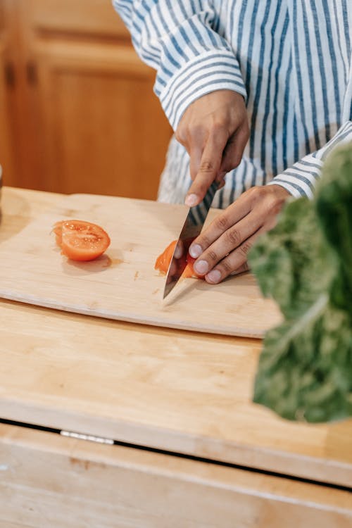 Crop ethnic man cutting fresh tomato while cooking at home
