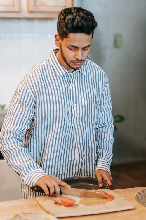 Young ethnic male in striped shirt with bowl and knife against chopping board with cut fresh tomato at home