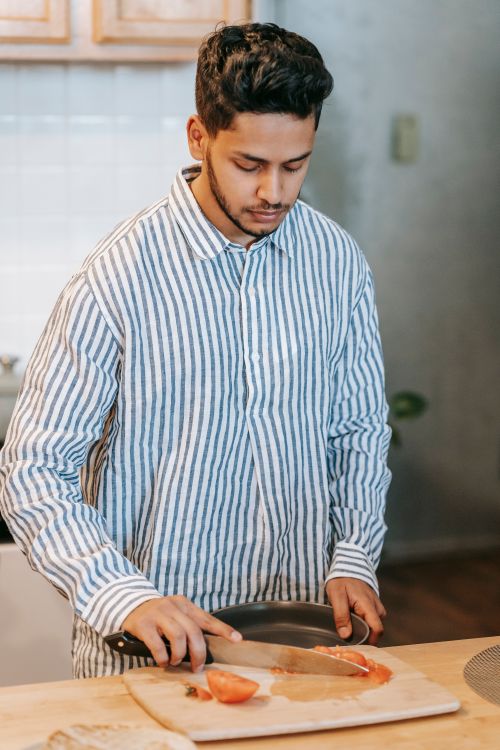 Free Young ethnic male in striped shirt with bowl and knife against chopping board with cut fresh tomato at home Stock Photo