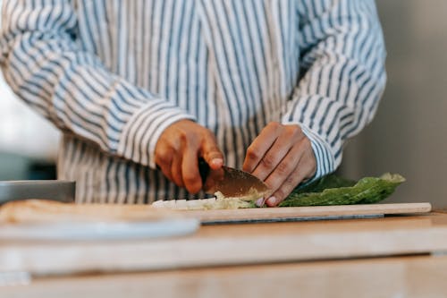 Crop anonymous male cook in striped shirt cutting fresh green lettuce leaves on wooden board while preparing dinner in kitchen