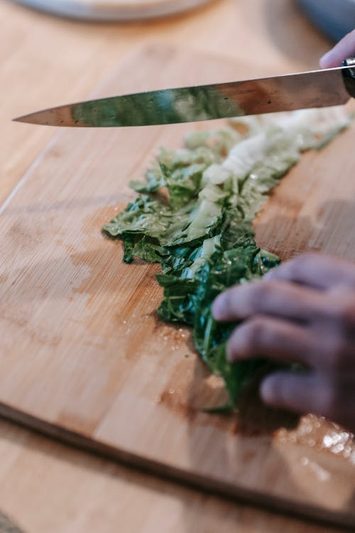 High angle of crop unrecognizable person cutting fresh lettuce with knife of wooden board during dinner preparation at home