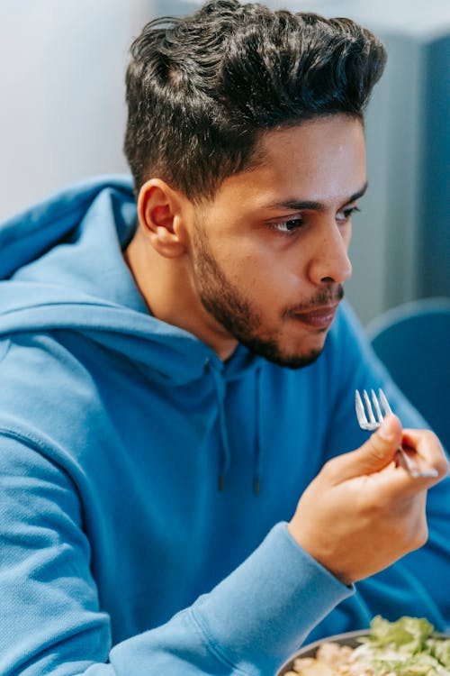 Free Young ethnic focused male in blue hoodie having meal with metal fork on blurred background Stock Photo