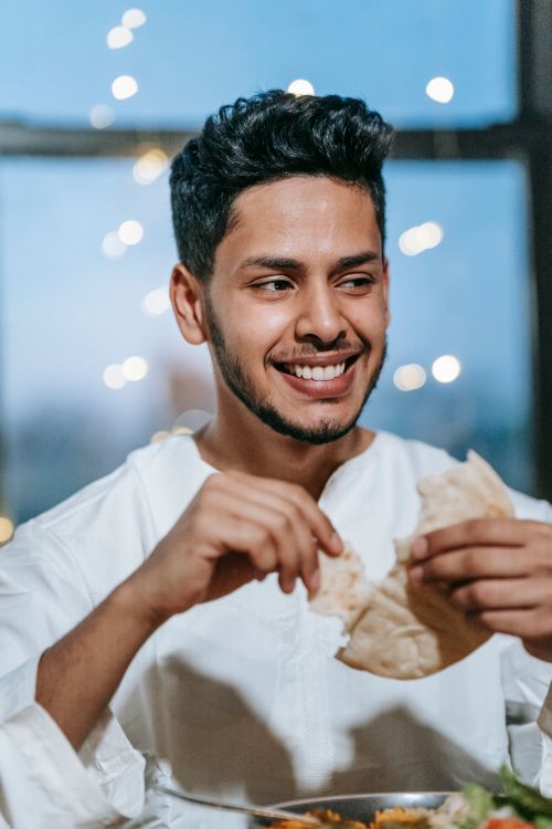 Free A man in a white thobe smiles while eating pita bread with string lights in the background, conveying a warm dining atmosphere. Stock Photo