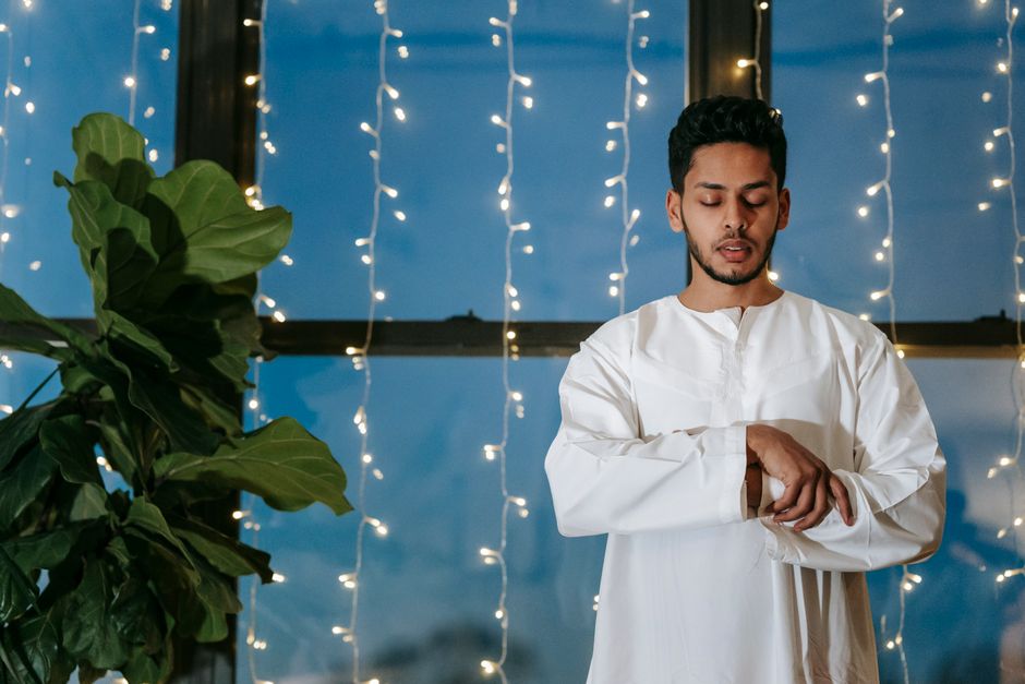 A man wearing traditional attire stands indoors near a window, illuminated by string lights, engaged in prayer.