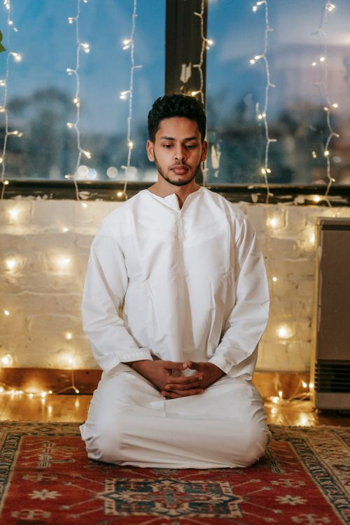 Free A man in traditional attire prays indoors, surrounded by warm string lights. Stock Photo