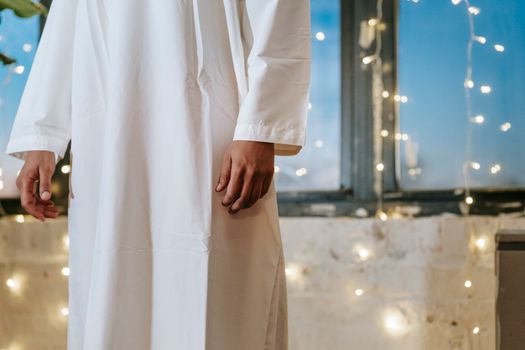 A man wearing a traditional white thobe stands indoors with string lights in the background.
