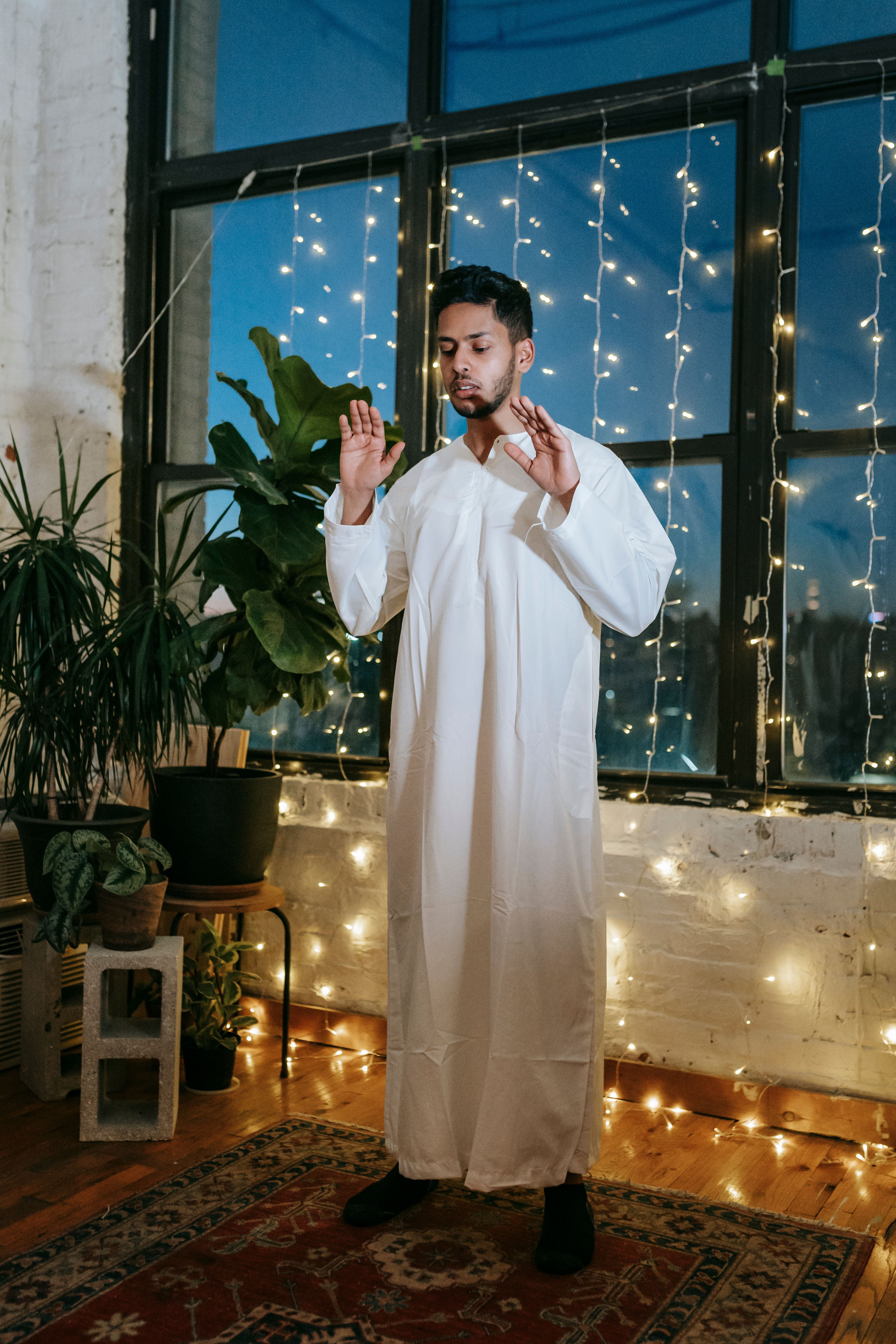 A man in traditional wear praying indoors with string lights enhancing the serene atmosphere.