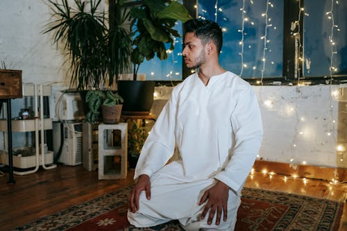 Pensive Indian male in white clothes standing on knees and looking away in room with garland in evening time