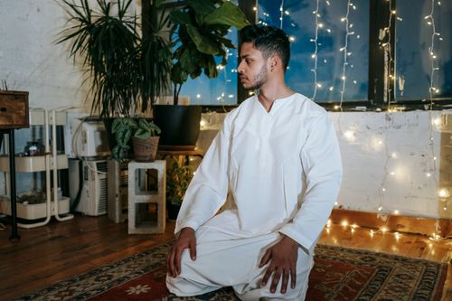 Free Pensive Indian male in white clothes standing on knees and looking away in room with garland in evening time Stock Photo