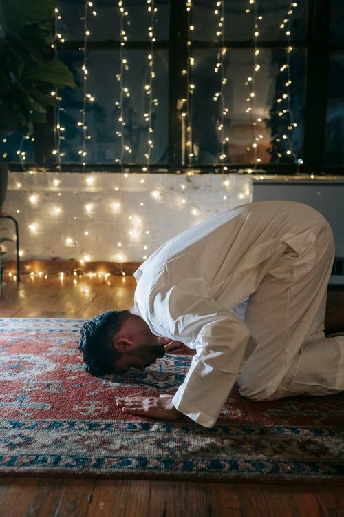 Free A Muslim man engaged in Islamic prayer at home, surrounded by glowing string lights and a rug. Stock Photo