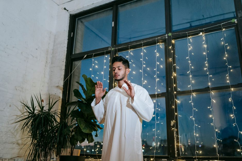 A man in traditional attire stands praying indoors by a window with string lights.