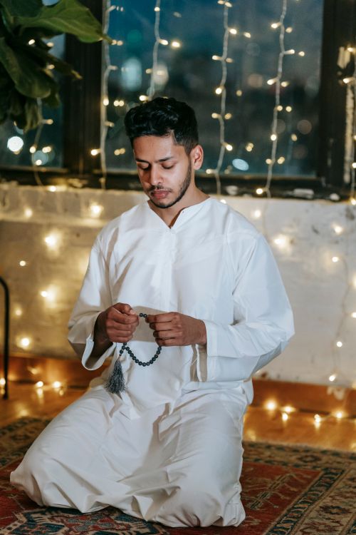 Free A man in traditional attire kneels in prayer indoors, holding beads with string lights in the background. Stock Photo