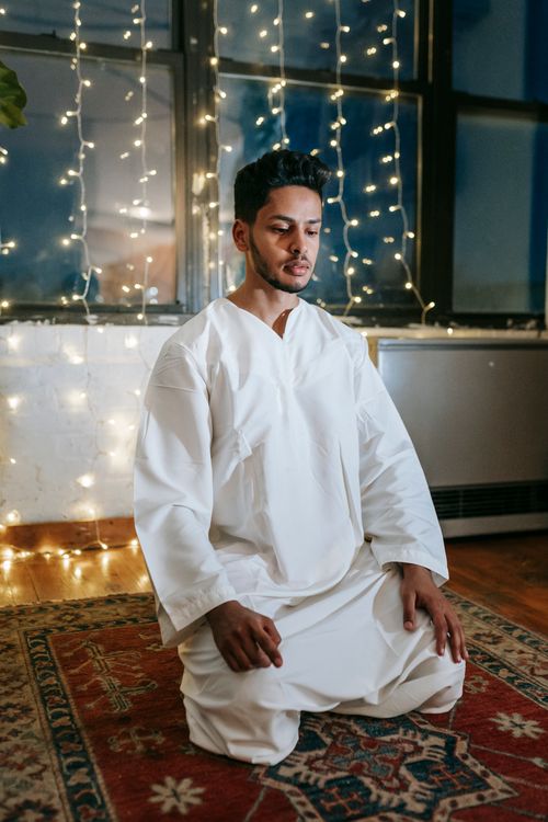 Free Man kneeling in prayer wearing a white thobe, surrounded by string lights in a cozy room. Stock Photo