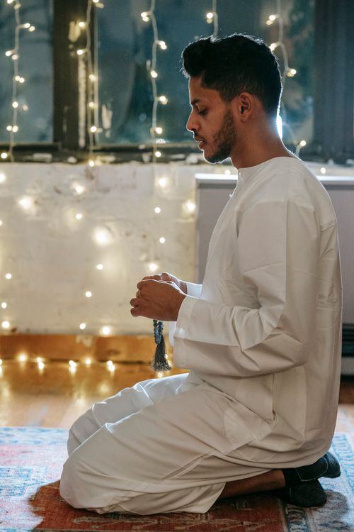 Free Side view of a Muslim man in traditional clothing praying indoors with string lights in the background. Stock Photo
