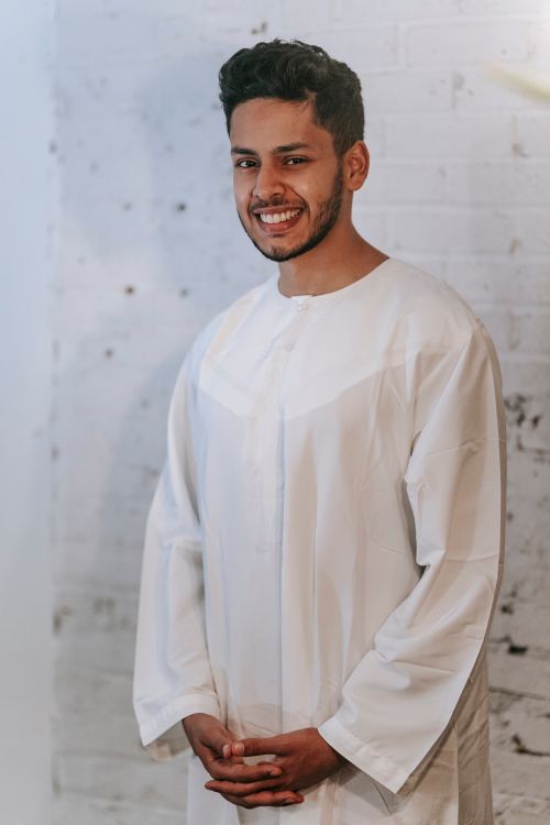 Free A happy man wearing a traditional Islamic thobe stands against a brick wall in an indoor setting. Stock Photo