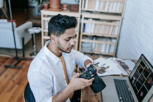 Side view of Indian male photographer in casual clothes watching photos on photo camera while sitting at table and browsing laptop at home in daytime