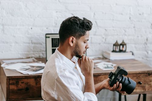 Free Side view of young ethnic guy in white shirt with professional photo camera near table with various photos and laptop with closed eyes in light apartment Stock Photo