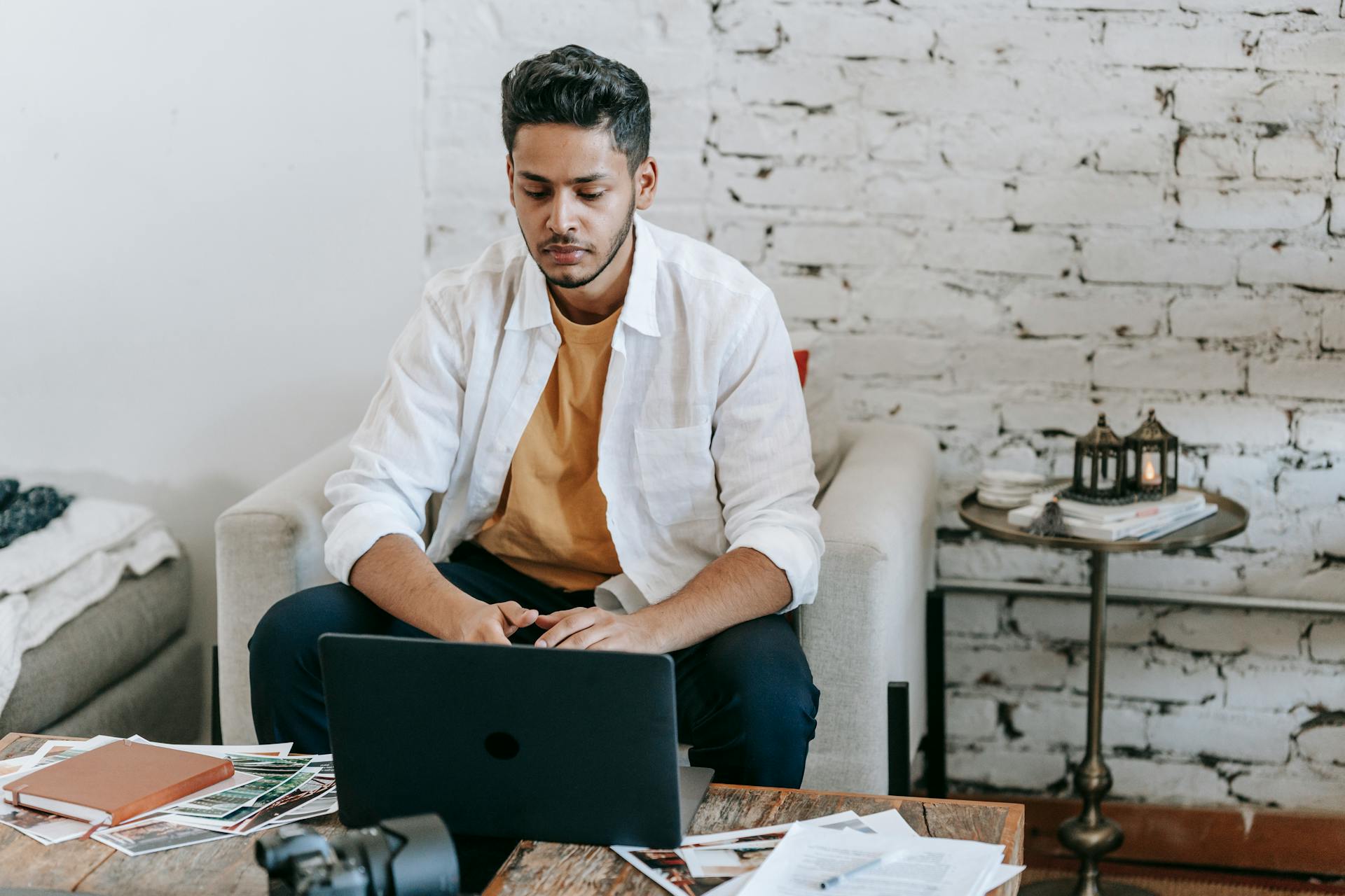 Creative young Hispanic male photographer sitting at table with photo camera and printed photos and working on laptop in modern loft style workplace at home