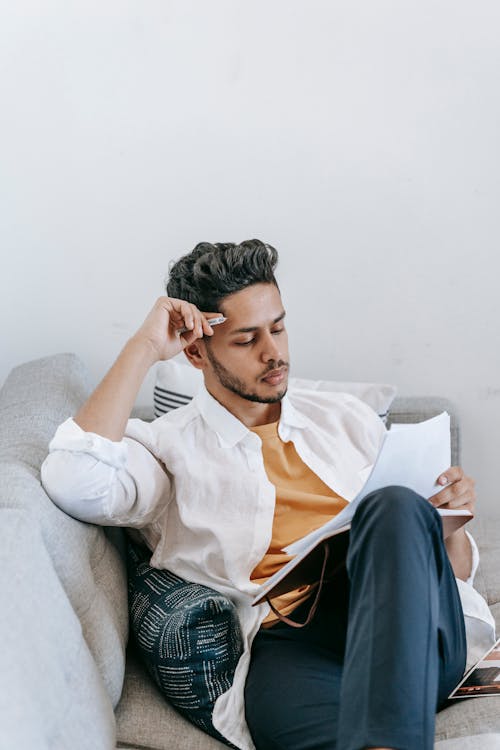 Focused young bearded Hispanic male entrepreneur in casual outfit with pen in hand sitting on sofa and reading papers while analyzing details of business project at home