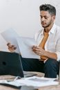 Serious young bearded Hispanic male journalist reviewing article on paper while working at table with laptop at workplace