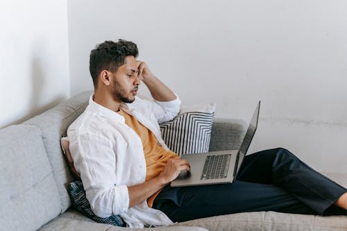 Side view of serious young Hispanic male remote employee sitting on sofa and using laptop while working distantly from home
