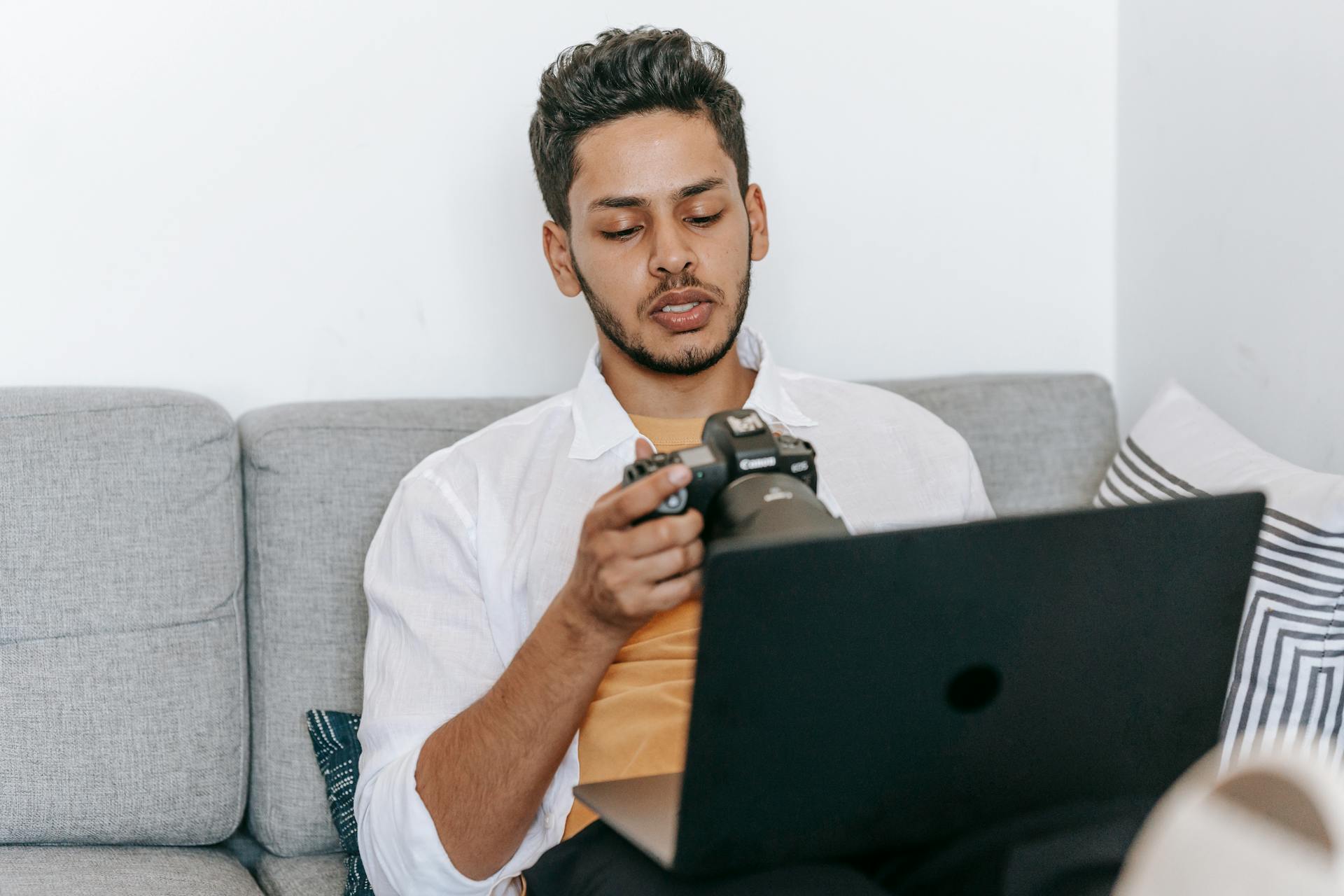 Young bearded ethnic male photographer checking pictures on photo camera while sitting with laptop on sofa and working on freelance project