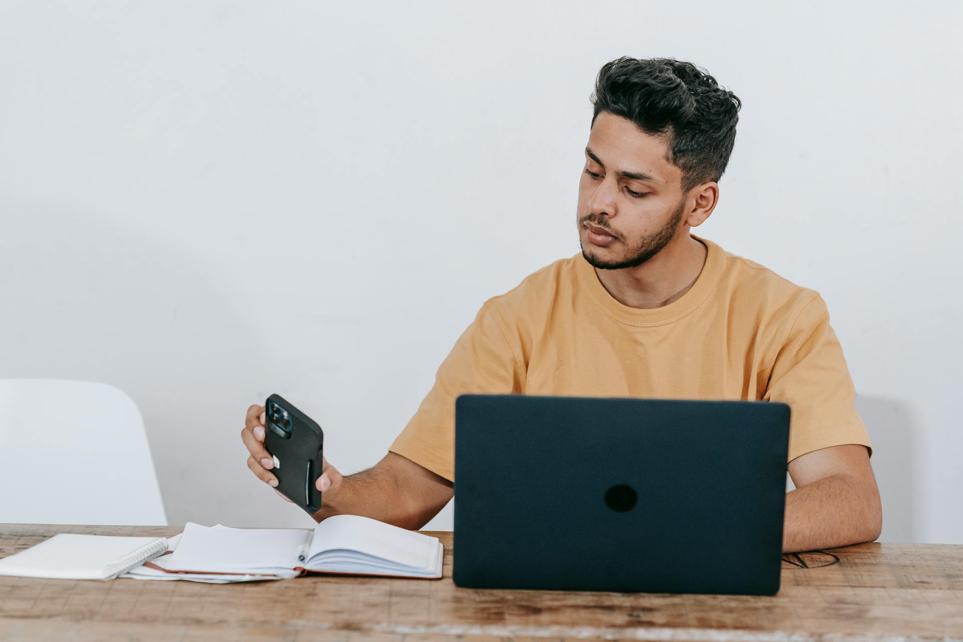 Serious young bearded Hispanic male entrepreneur in casual clothes checking notifications on mobile phone while working at table with laptop and notebook at workplace