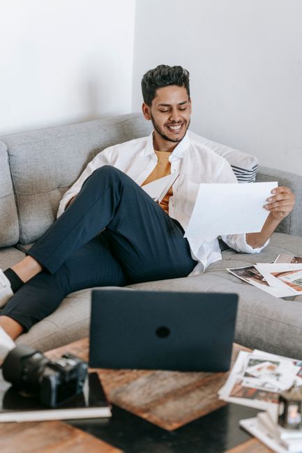 Positive young Hispanic male photographer checking printed photos and smiling satisfied with quality while working on sofa near laptop and photo camera at home Positive young Hispanic male photographer checking printed photos and smiling satisfied with quality while working on sofa near laptop and photo camera at home