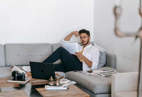 Free Creative young Hispanic male photographer with papers and pen in hand thinking about new project while sitting on sofa near laptop and photo camera at home Stock Photo