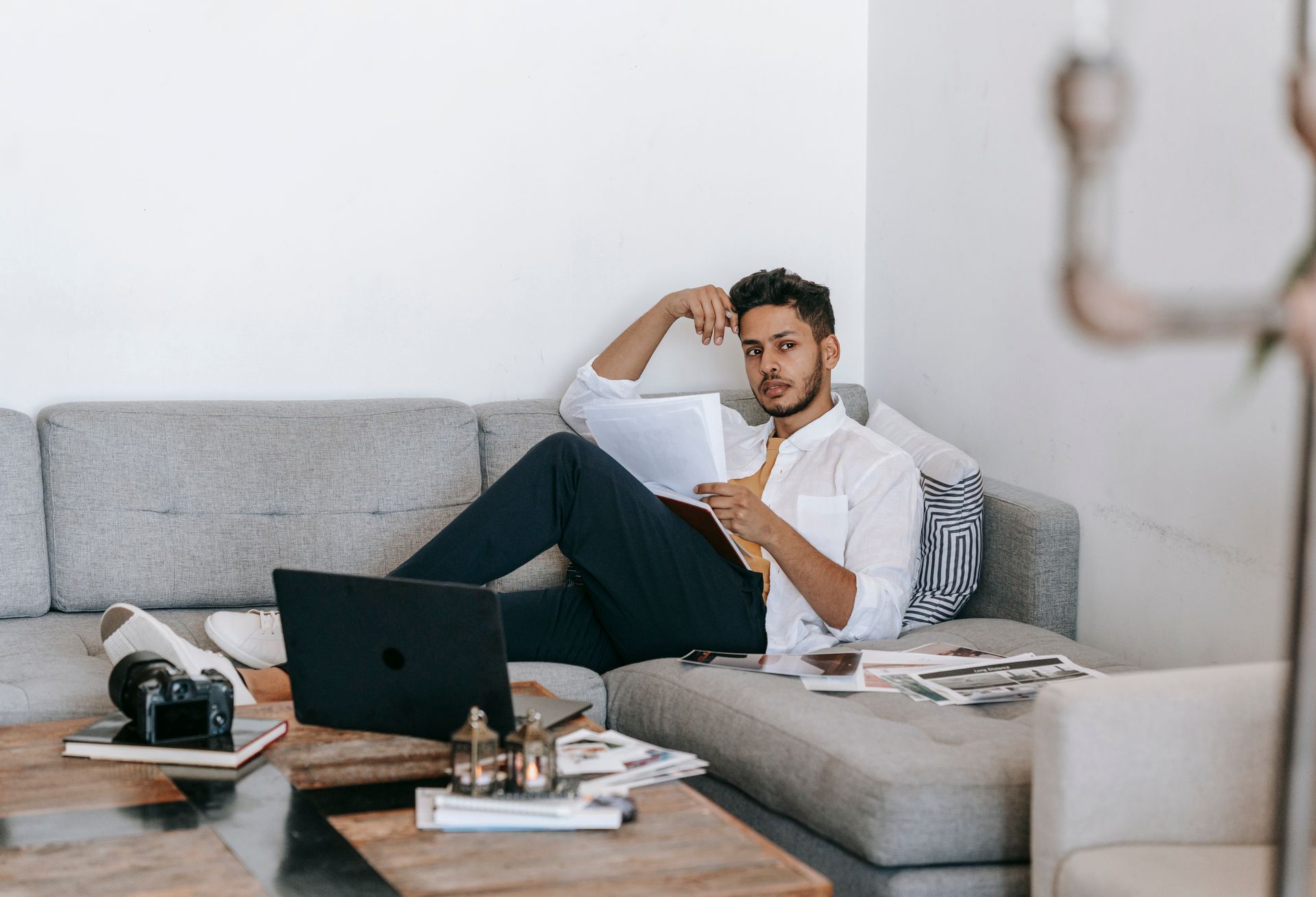 Creative young Hispanic male photographer with papers and pen in hand thinking about new project while sitting on sofa near laptop and photo camera at home