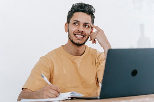 Smiling young bearded Hispanic male entrepreneur thinking over new ideas for startup project and looking away dreamily while working at table with laptop and taking notes in notebook