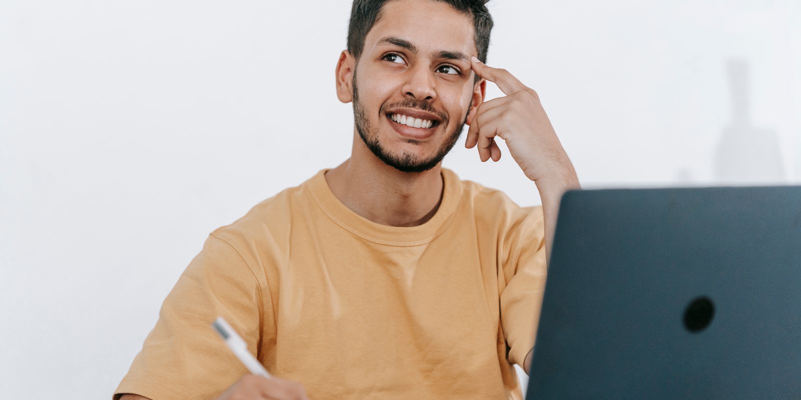 Smiling young bearded Hispanic male entrepreneur thinking over new ideas for startup project and looking away dreamily while working at tabl