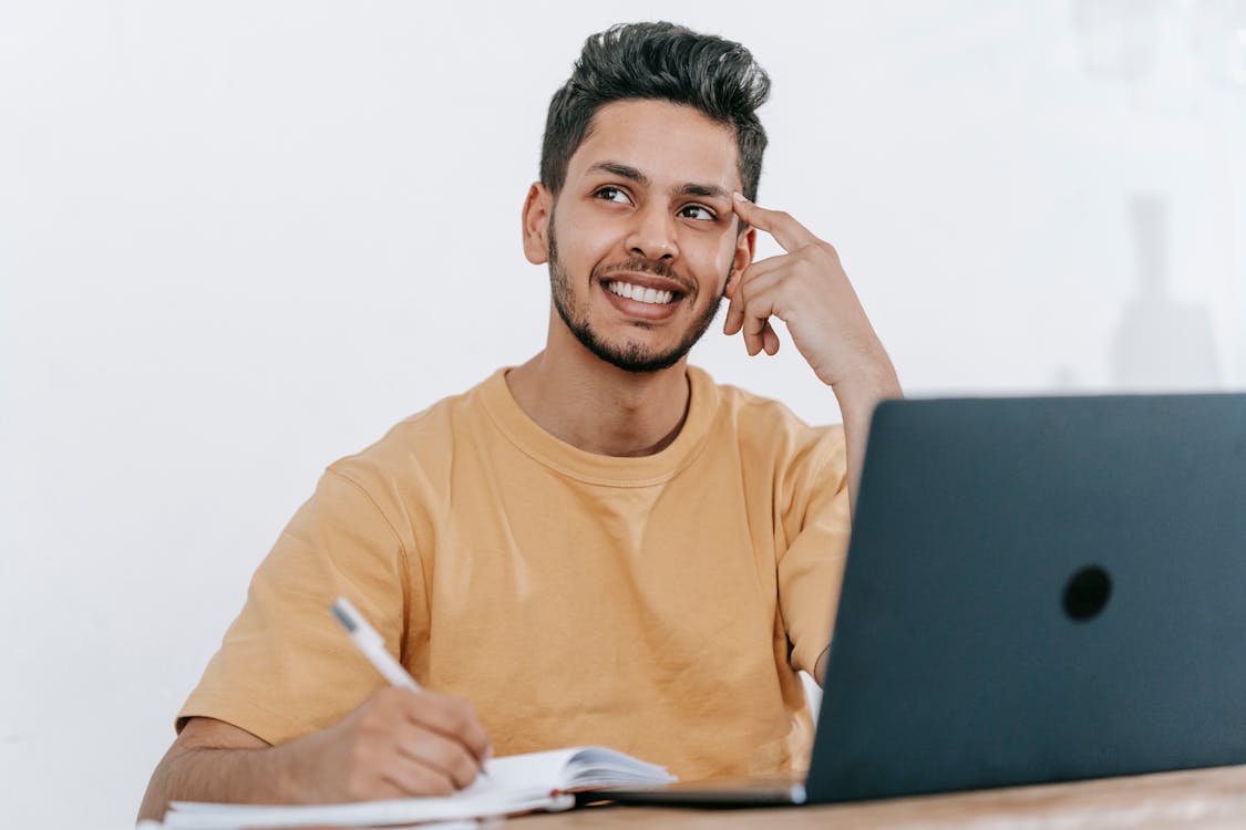 Free Smiling young bearded Hispanic male entrepreneur thinking over new ideas for startup project and looking away dreamily while working at table with laptop and taking notes in notebook Stock Photo