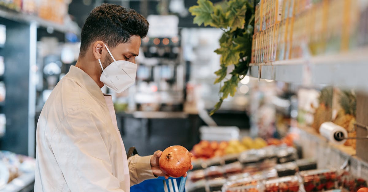Side view of young Hispanic man in protective mask choosing fresh pomegranate from stall in supermarket during coronavirus pandemic