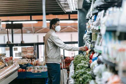 Free Side view of young Hispanic male buyer in protective mask with eco friendly cotton bag taking fresh vegetables from shelf in local grocery store during coronavirus pandemic Stock Photo