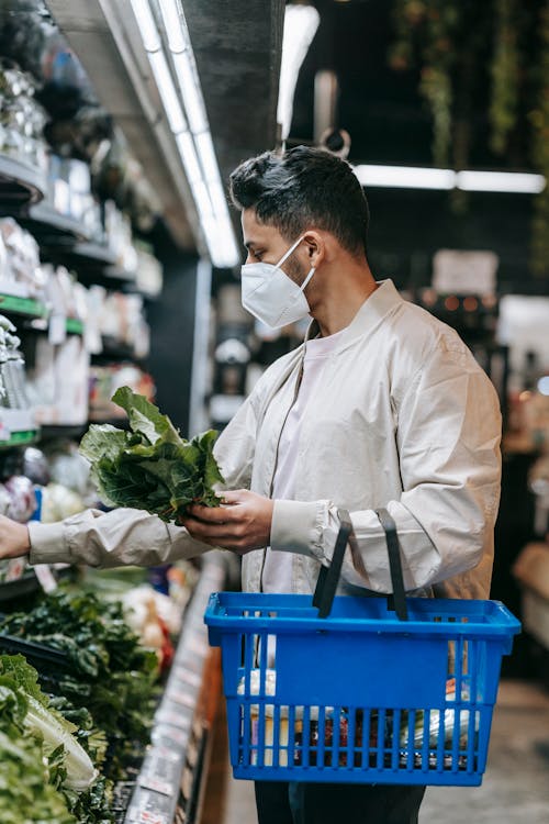 Side view of young ethnic male buyer in protective mask with shopping basket picking fresh green leaves from shelf in supermarket while doing purchases during coronavirus pandemic