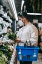 Side view of young ethnic male buyer in protective mask with shopping basket picking fresh green leaves from shelf in supermarket while doing purchases during coronavirus pandemic
