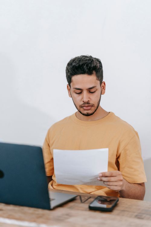 Free Serious young bearded Hispanic businessman in casual outfit reading contract while sitting at table with laptop in office Stock Photo