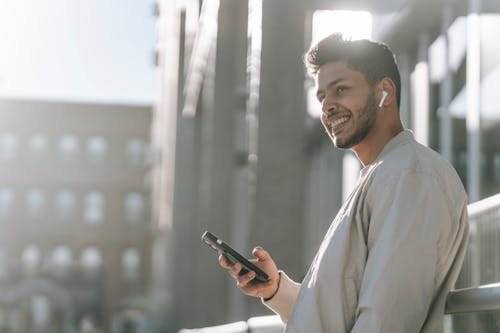 Side view of cheerful young bearded Hispanic male in TWS earbuds using mobile phone while standing near modern urban building in sunny day