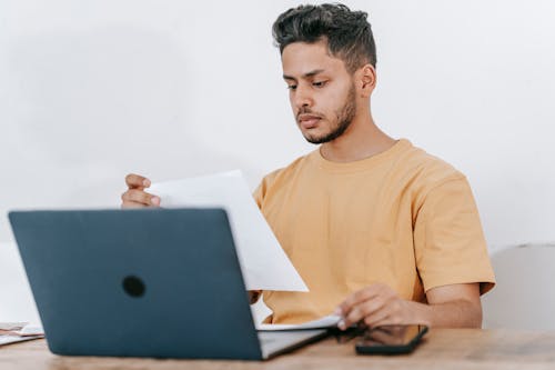 Young bearded Hispanic male university student reading information on paper while studying online via laptop