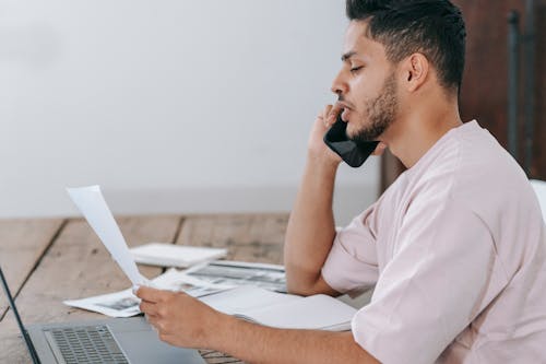 Side view of young Hispanic male remote employee discussing documents during phone call while working online with laptop and papers in home office
