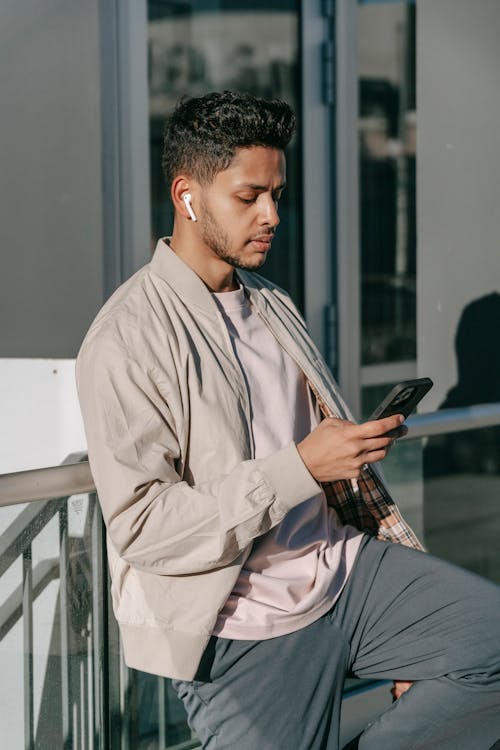 Free Serious young bearded Hispanic male in casual clothes listening to audio message via mobile phone and TWS earbuds while standing near modern city building Stock Photo