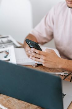 Crop unrecognizable male text messaging on cellphone while sitting at table with netbook and notebook during work in light room