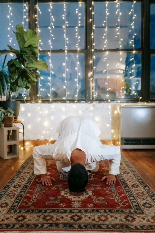 Free A man in traditional wear kneels on a prayer rug indoors, surrounded by string lights, embodying a serene religious practice. Stock Photo
