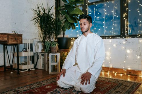 Free A young Muslim man kneels in prayer, surrounded by string lights indoors, creating a serene atmosphere. Stock Photo