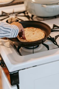 A hand cooking pita bread on a stovetop with a frying pan in a cozy kitchen setting.